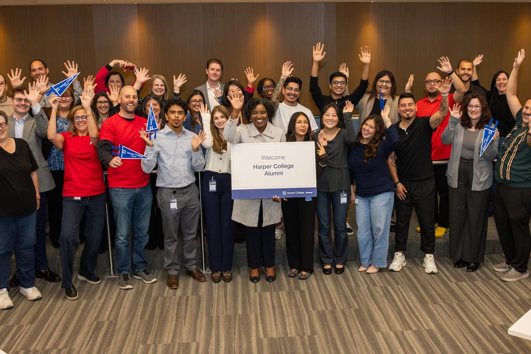 A large group of Harper College alumni and staff smiling and waving together during the Harper @ Work event at the Zurich Center.
