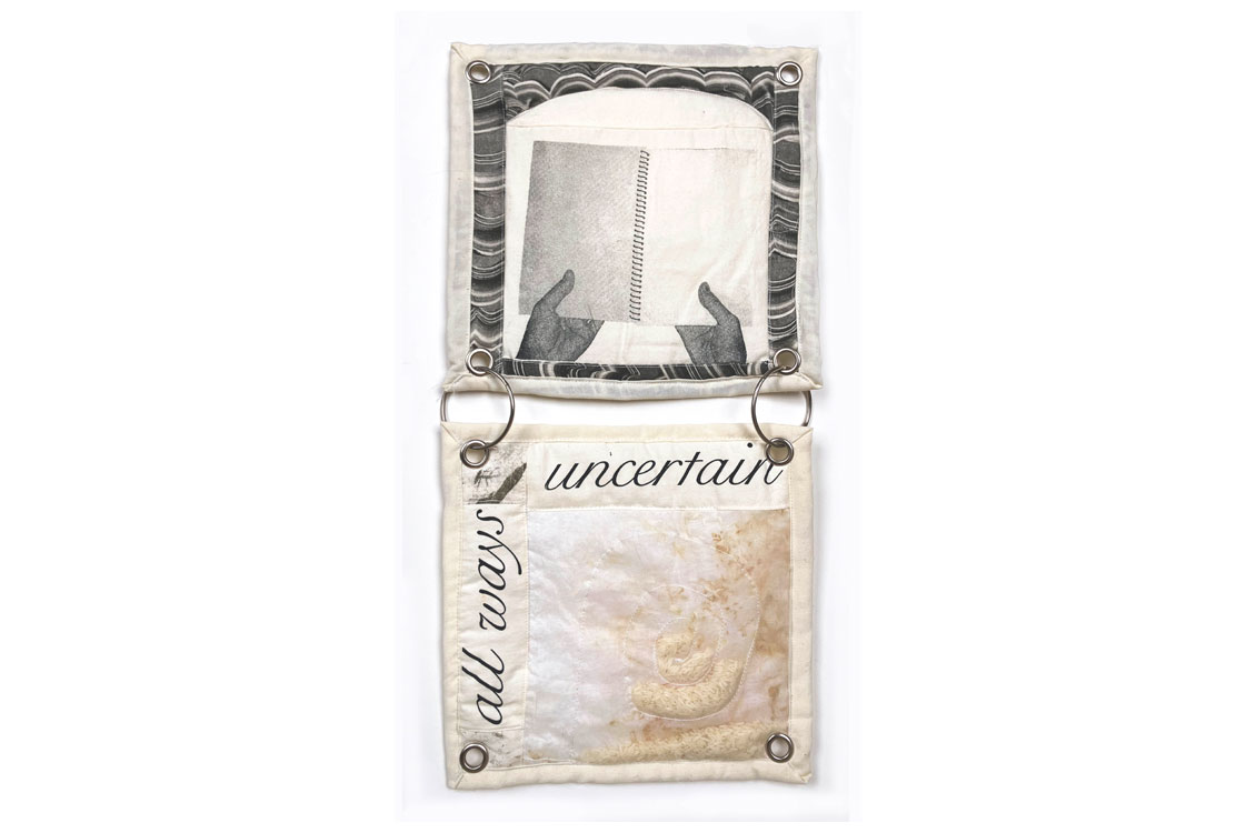 two fabric squares joined together with binder rings; the top square is a black and white image of hands holding a book and the bottom is an abstract piece with the words 'all ways uncertain' written on it