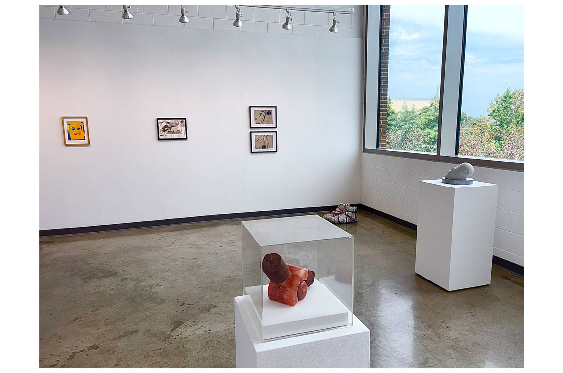 Gallery wall with four framed artworks; a ceramic sculpture in a display case and a stone sculpture on pedestals nearby.