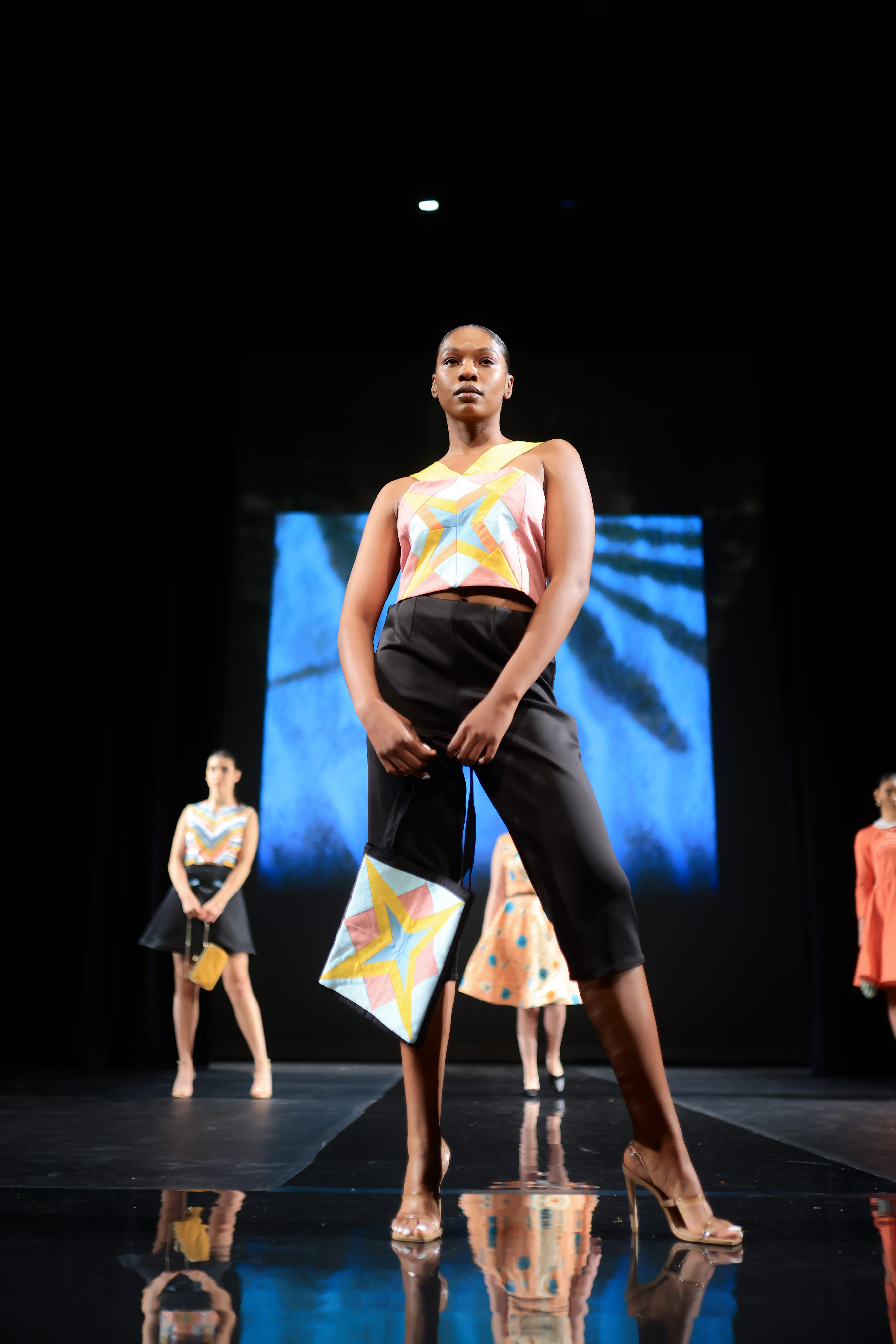 A model strikes a pose in stylish black capri slacks, transparent heels, and a vibrant sleeveless top adorned with a colorful pattern. Completing the look with a coordinating handbag.