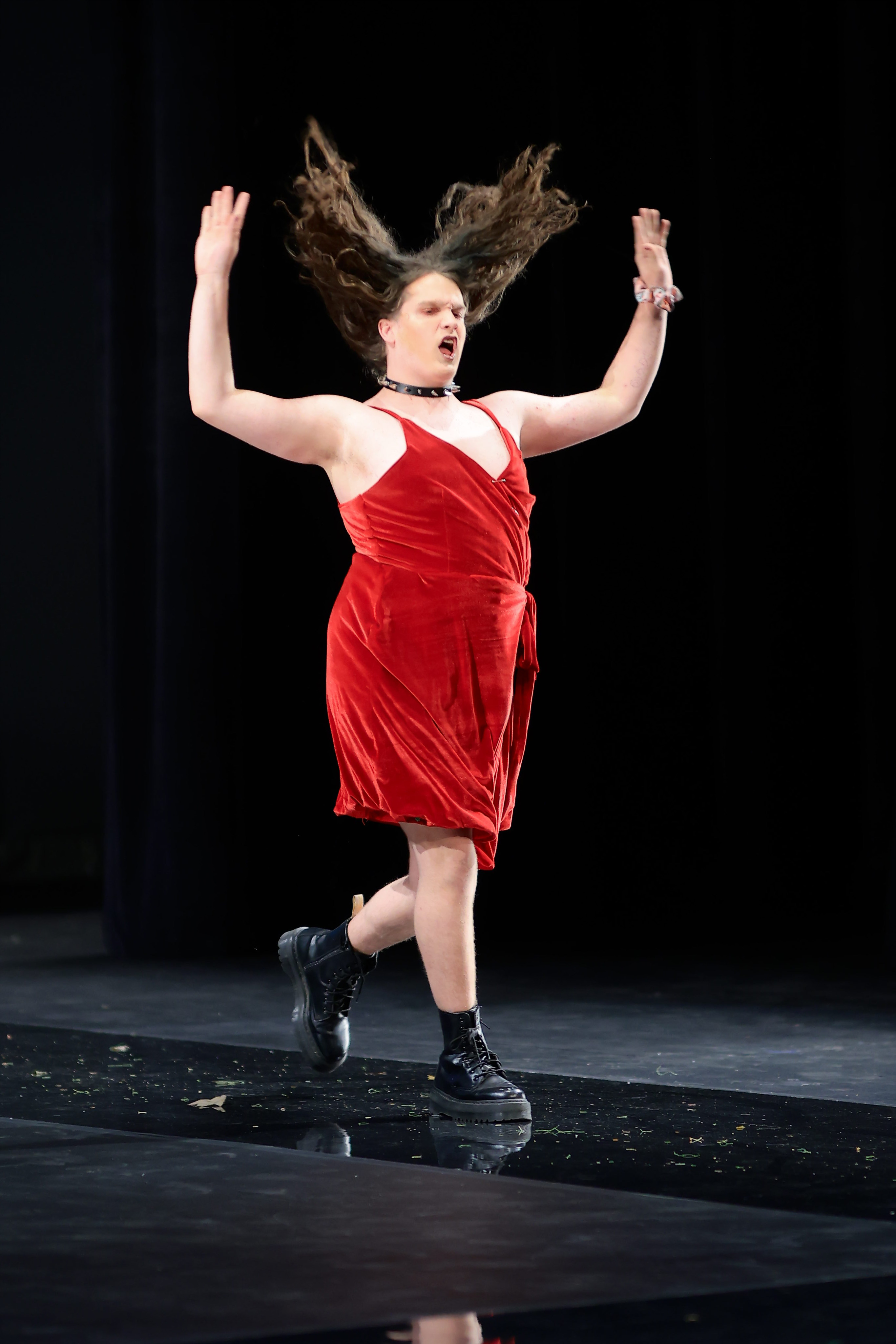 A model confidently walks the runway, flipping their hair, in a red sleeveless wrap dress paired with edgy black combat boots. They add a touch of attitude with a black studded choker and a pop of color with a vibrant scrunchie on their left wrist.