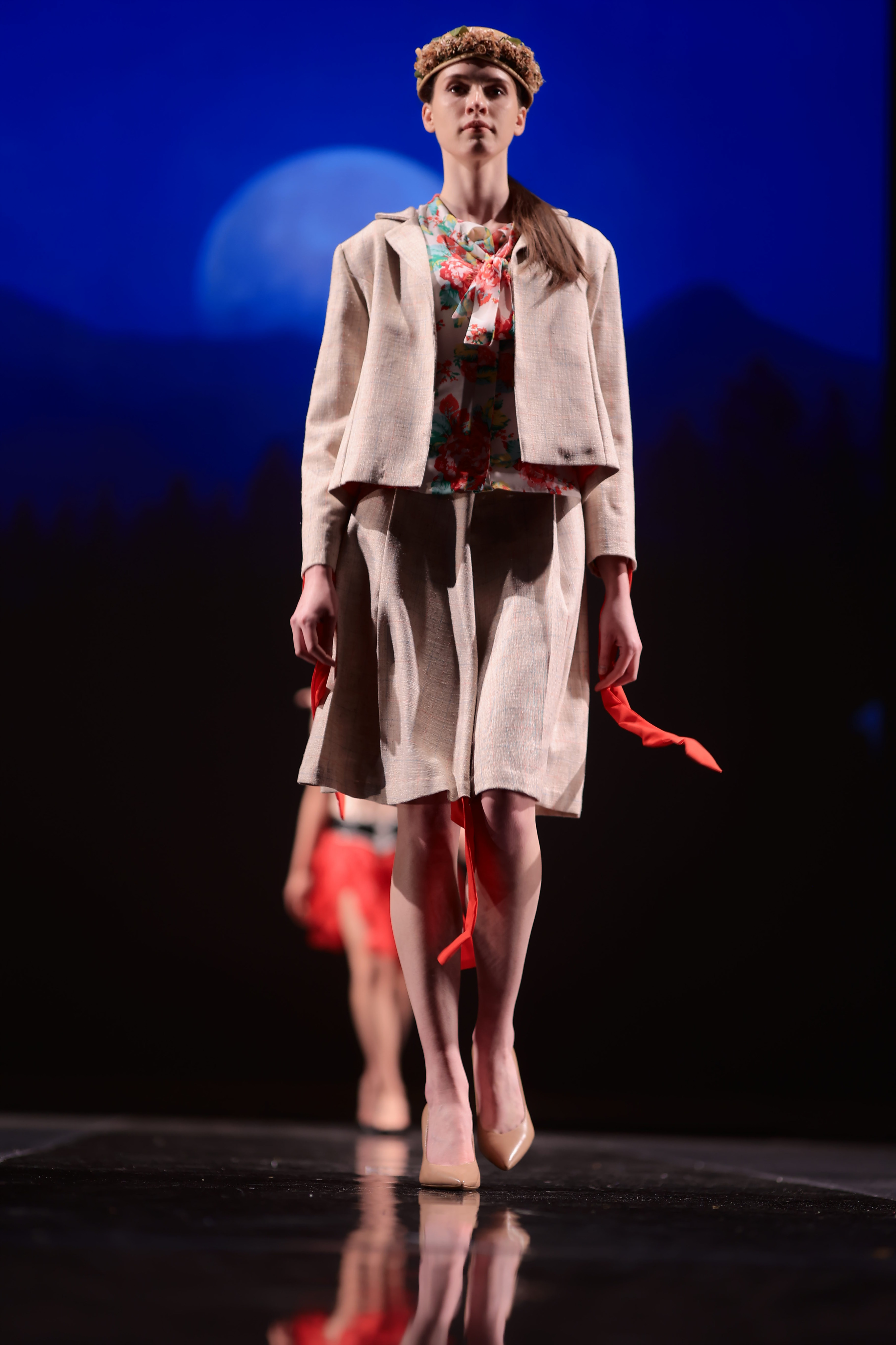 A model struts down the runway in a sophisticated beige dress suit paired with a colorful patterned top. Completing the look with a dark brown hat and beige heels.