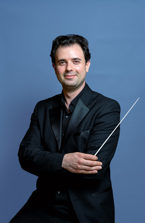 Man in a suit standing with arms crossed, holding a conducting baton