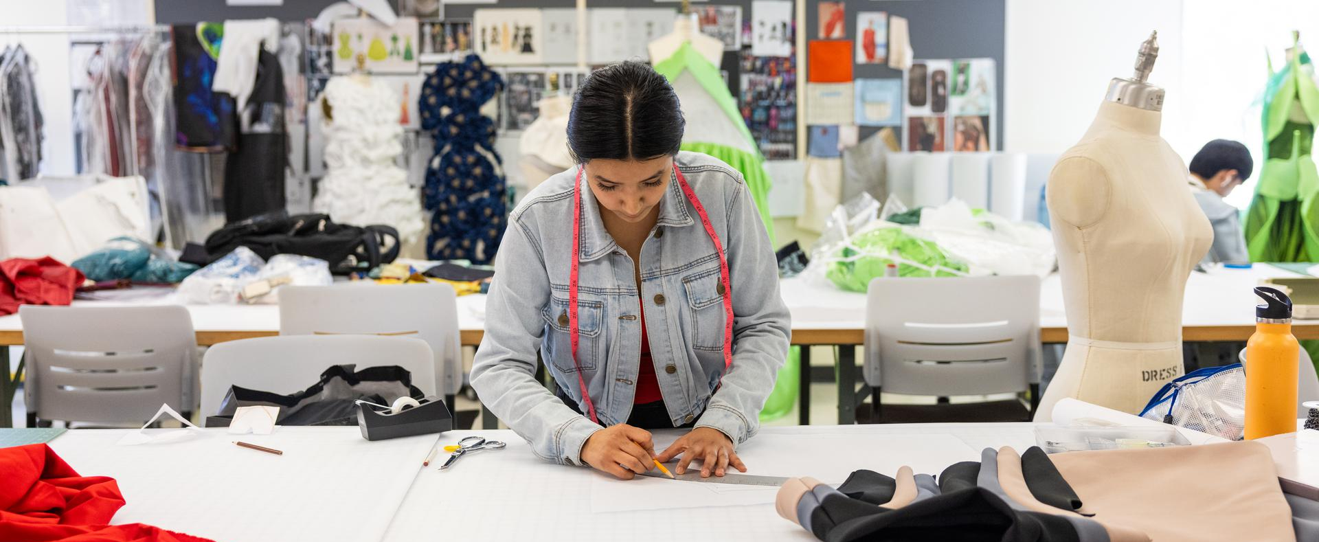 Student working on a fashion design pattern in a classroom with fabric, tools, and mannequins.