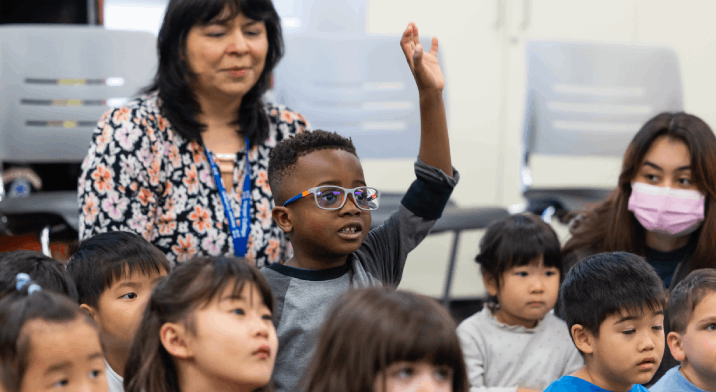 Pre-school students sit in a group while one raises his hand for a question.