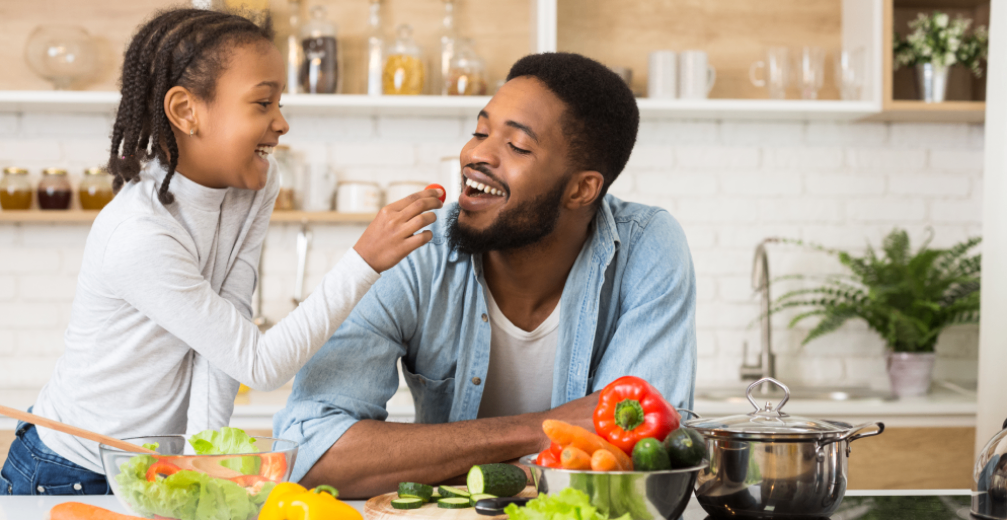 A girl feeds a cherry tomato to a smiling man in a kitchen.