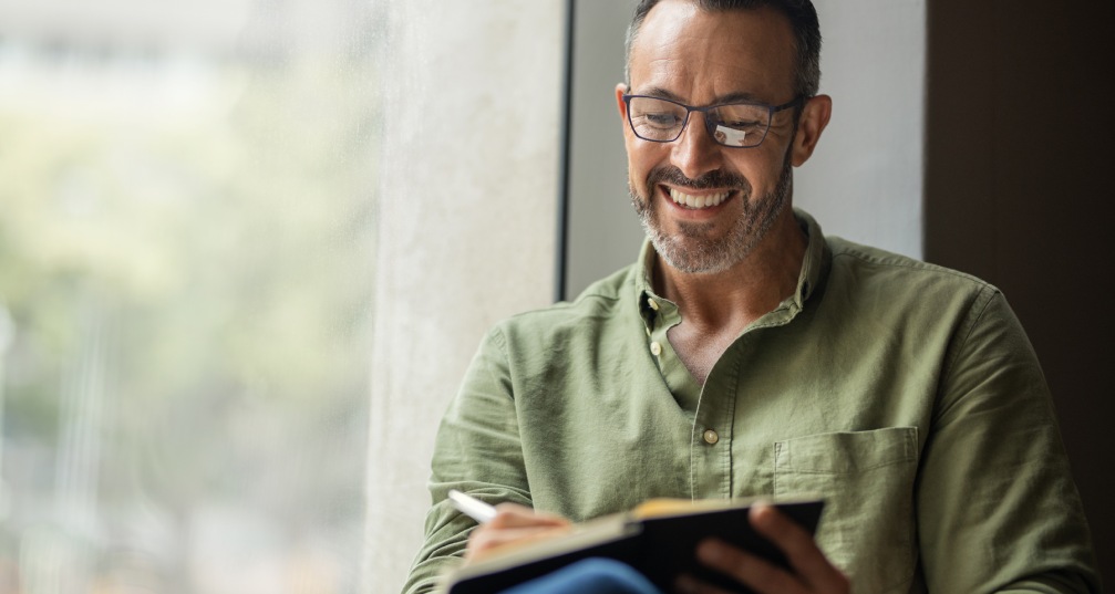 a smiling man sitting by a window and writing in a journal