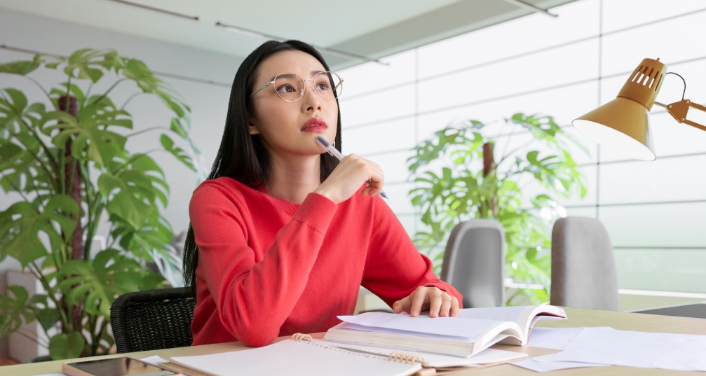 a woman sits at her desk, looking up thoughtfully while working on a budget