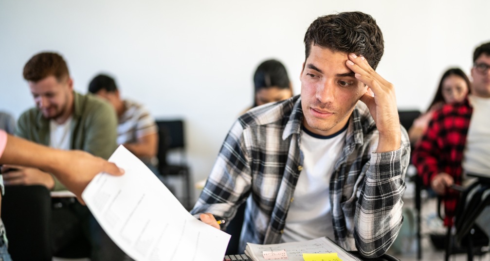 a young man at a desk looking upset as he is handed back a paper containing a disappointing grade