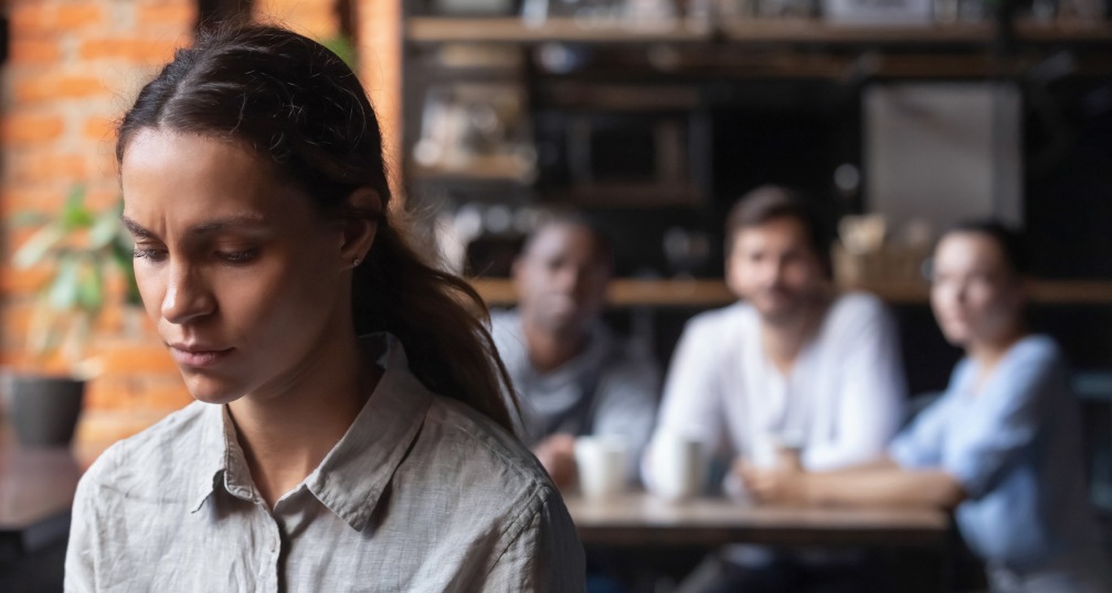 a woman in focus sits alone at a table looking downcast, while 3 people out of focus sit at a table further back and watch her