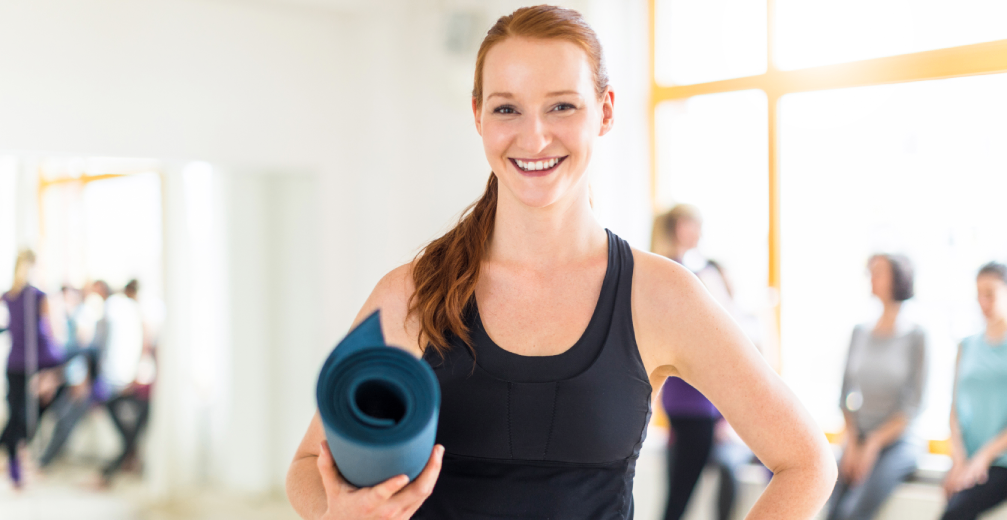 Smiling woman holding a yoga mat in a fitness studio.