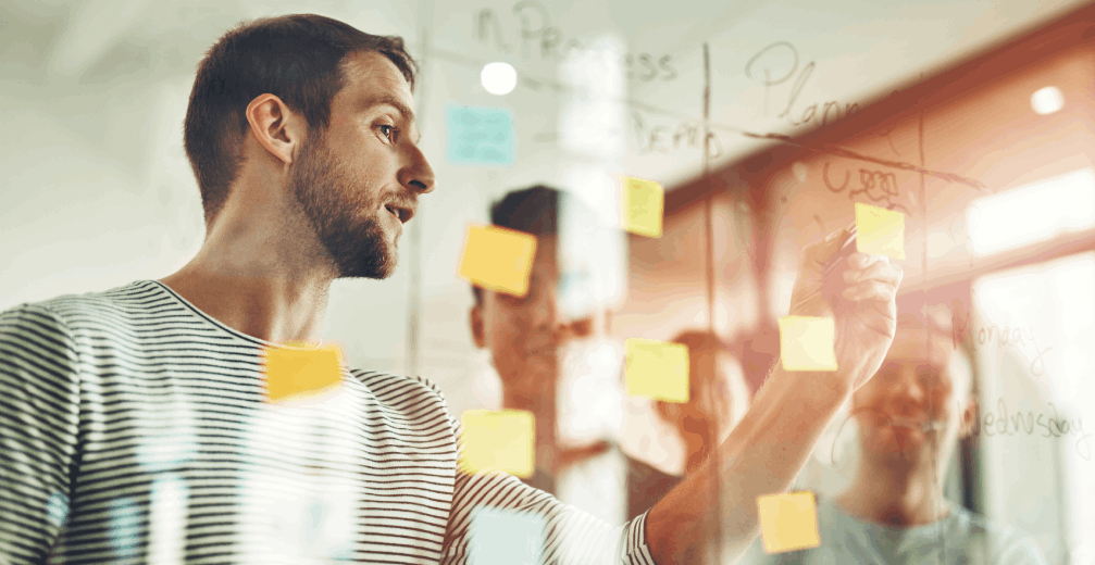 Man writing on a glass board with sticky notes.