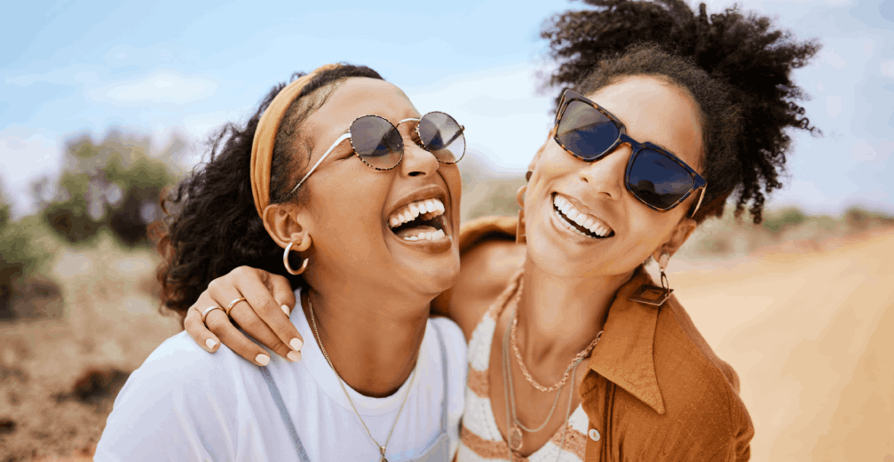 Two friends wearing sunglasses laughing together outdoors.