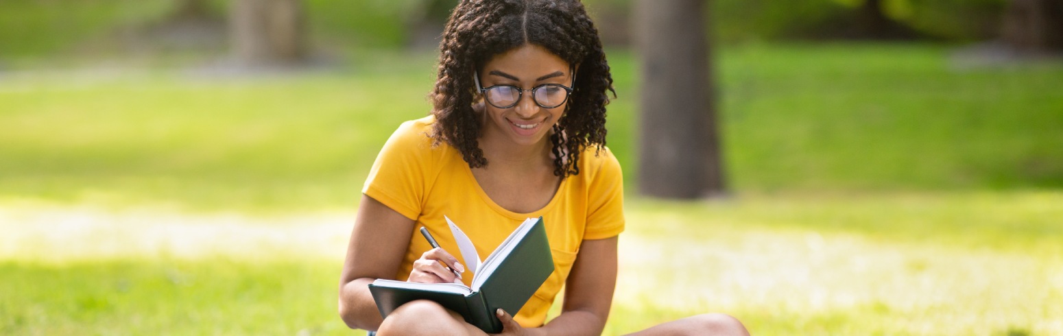 Young woman writing in a journal with her laptop in a grassy park.