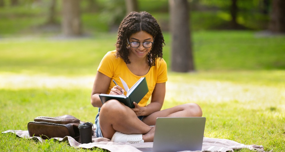 Young woman writing in a journal with her laptop in a grassy park.