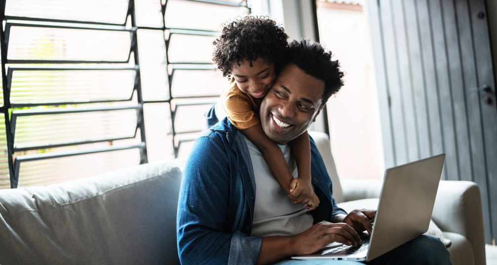 A man sitting on the couch and using a laptop while his son hugs him from behind