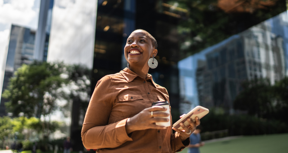 a woman holding a coffee cup in a downtown setting looks up from her phone with a hopeful expression