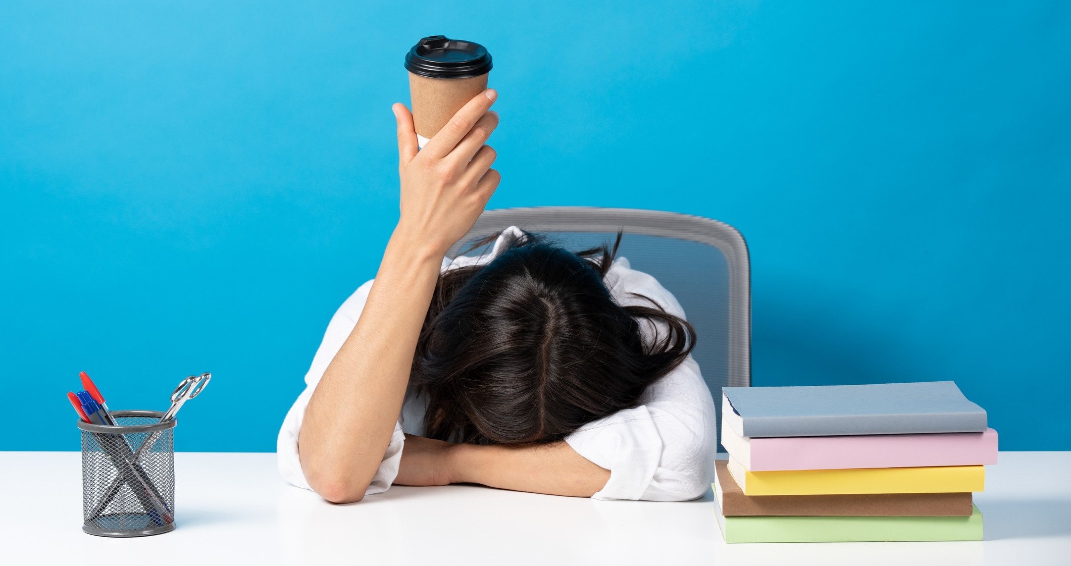 stressed woman holding up a coffee cup, resting her head on a desk with a cup of pens and a stack of books on top, isolated on blue background