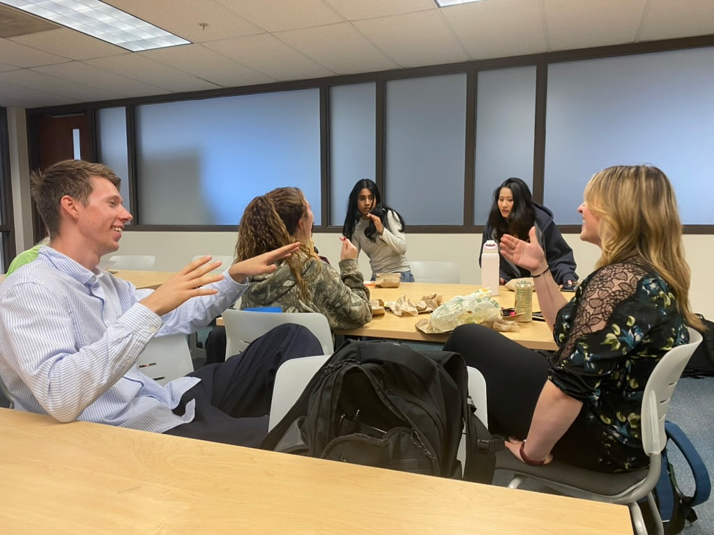 Harper College Business Club members interacting informally during a meeting, with students seated at tables and talking