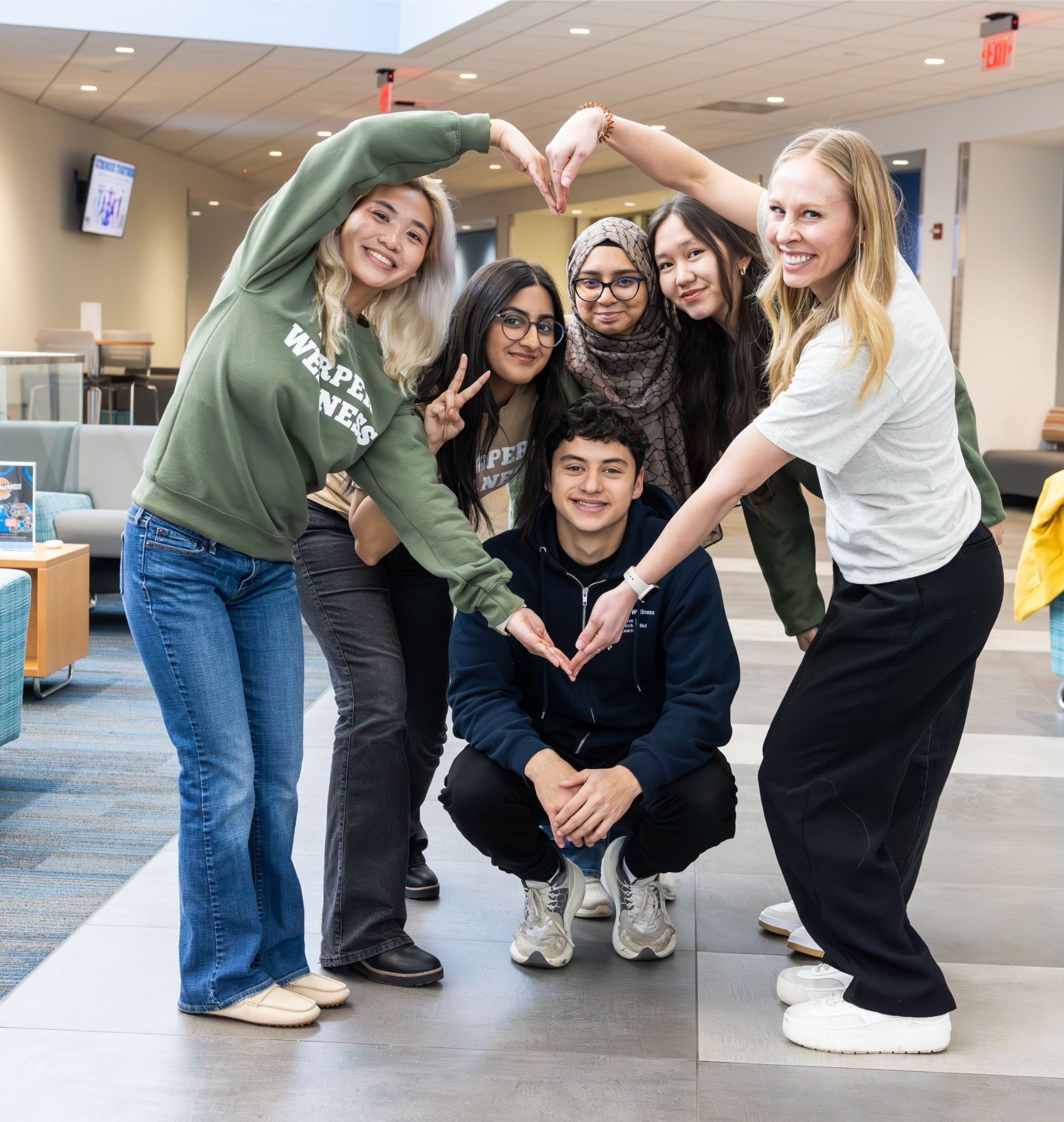 Six adults pose in a lobby, forming heart shapes with their arms around a crouching person in the center.